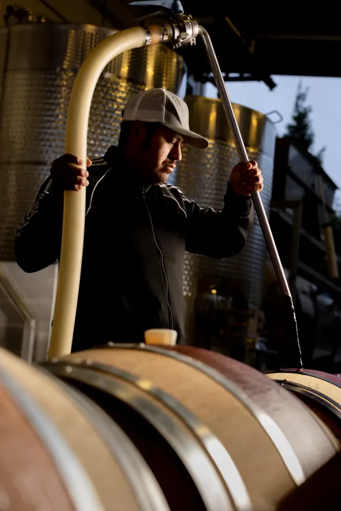 A person in a cap works with barrels and hoses in a winery or brewery setting.