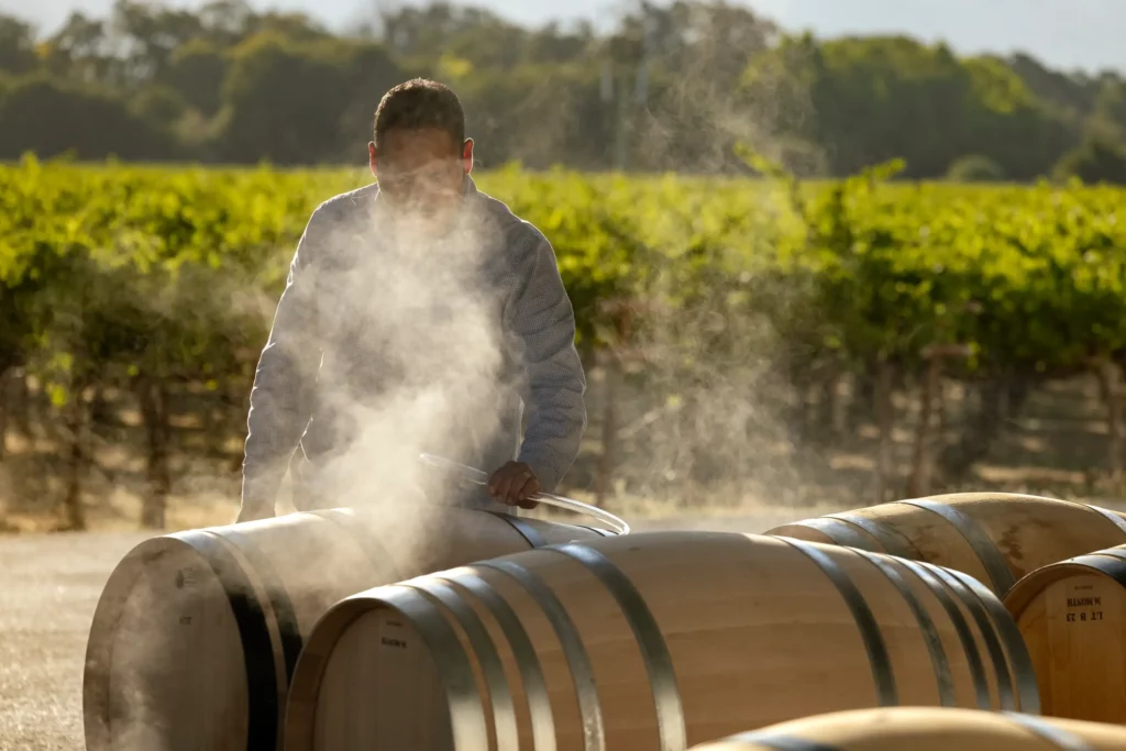 Person working with steaming wine barrels in a vineyard setting.