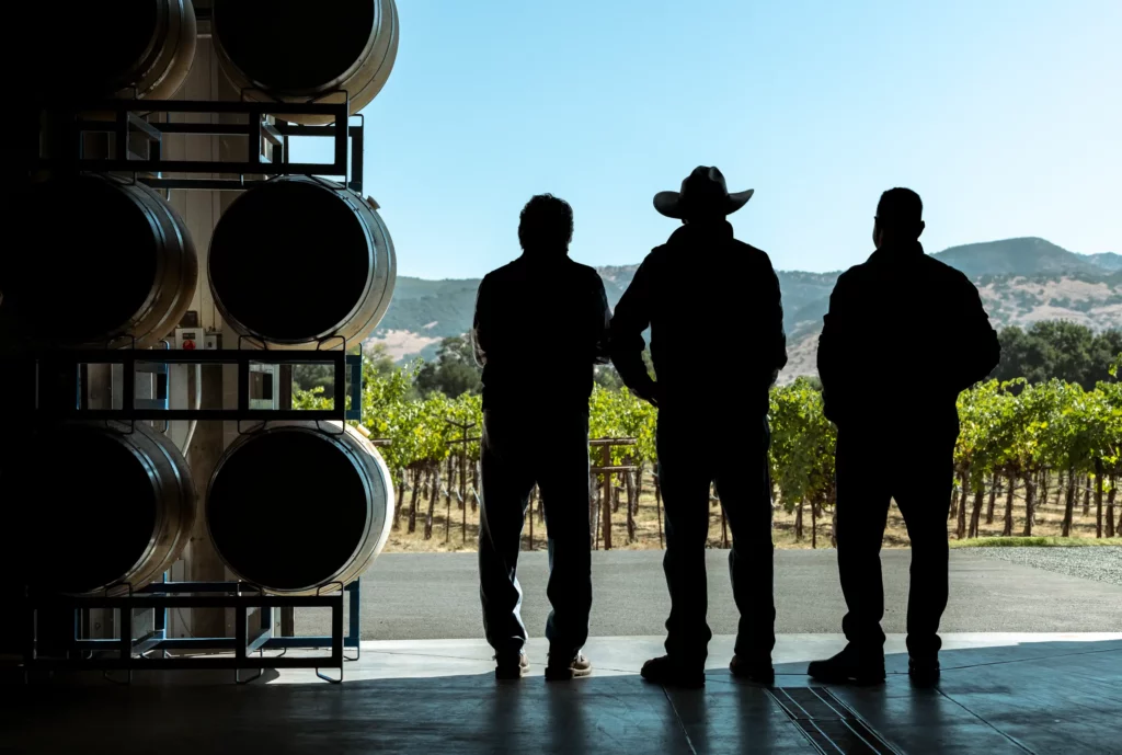 Three people standing in a winery, silhouetted against sunlight. Large barrels are on the left, and vineyards stretch into the distance with hills on the horizon.