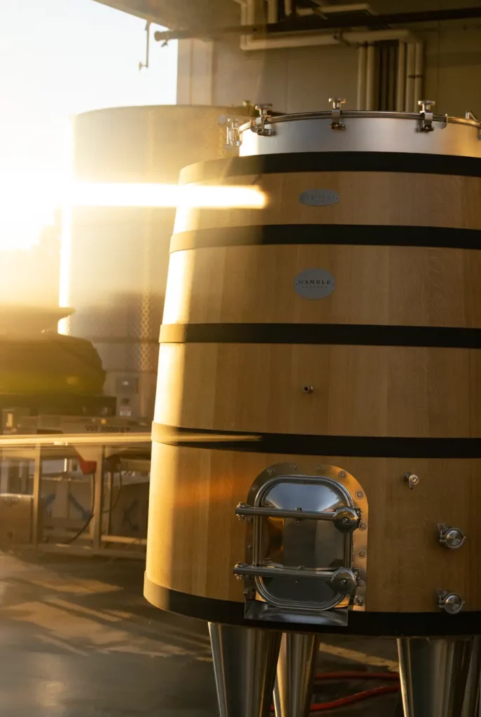 A large wooden fermentation tank with metal fittings is illuminated by sunlight in a warehouse setting.