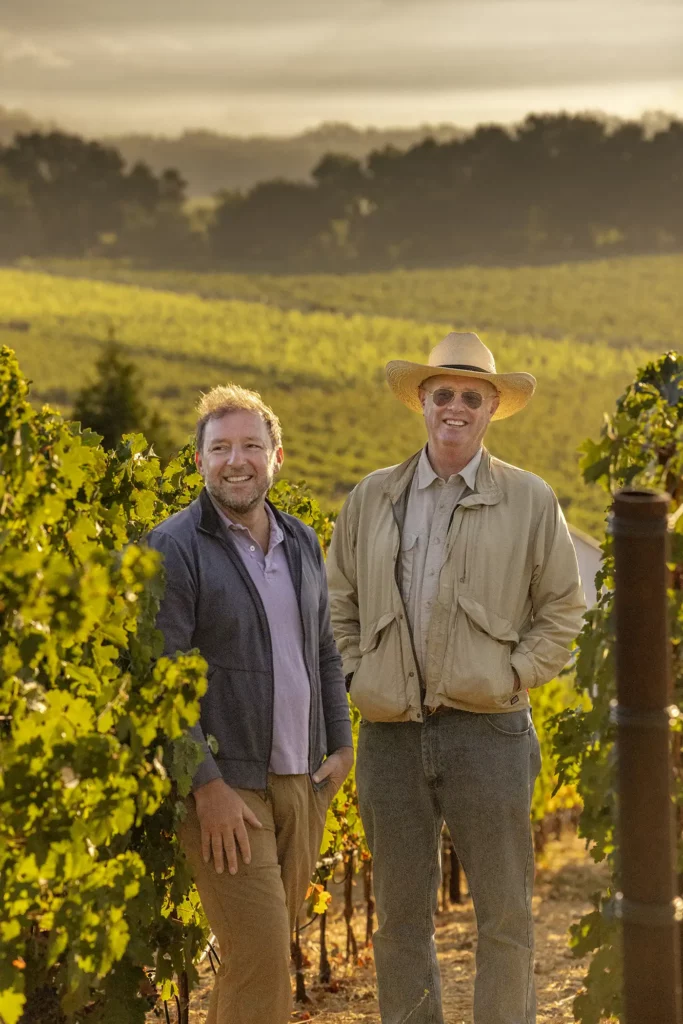 Two men stand in a vineyard, one wearing a hat and sunglasses. Lush grapevines and hills are in the background under a hazy sky.