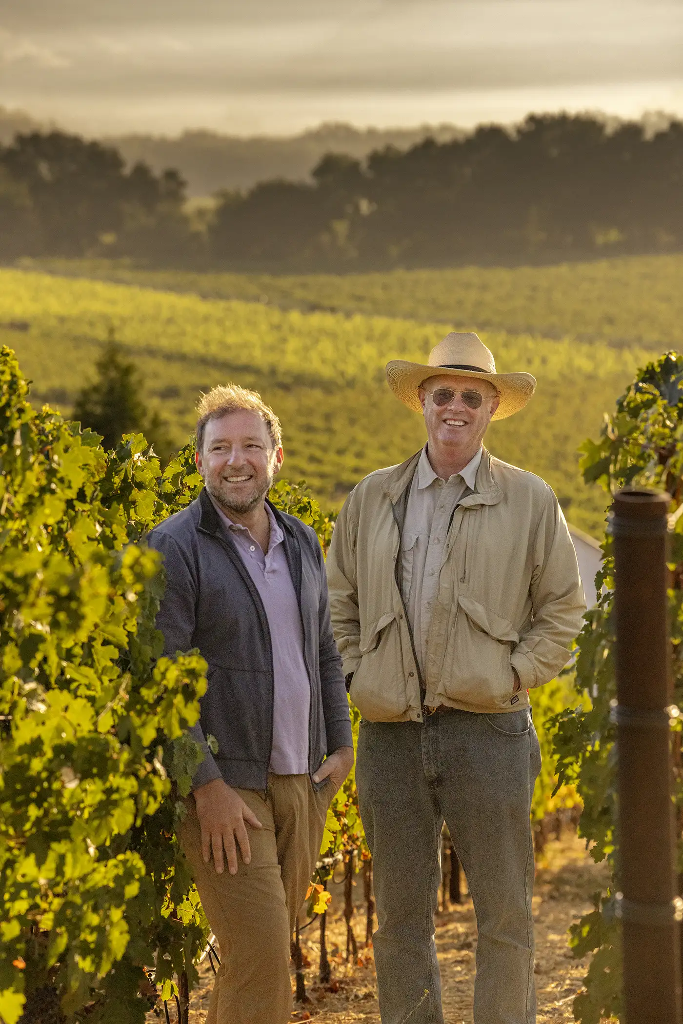 Two men stand in a vineyard, one wearing a hat and sunglasses. Lush grapevines and hills are in the background under a hazy sky.